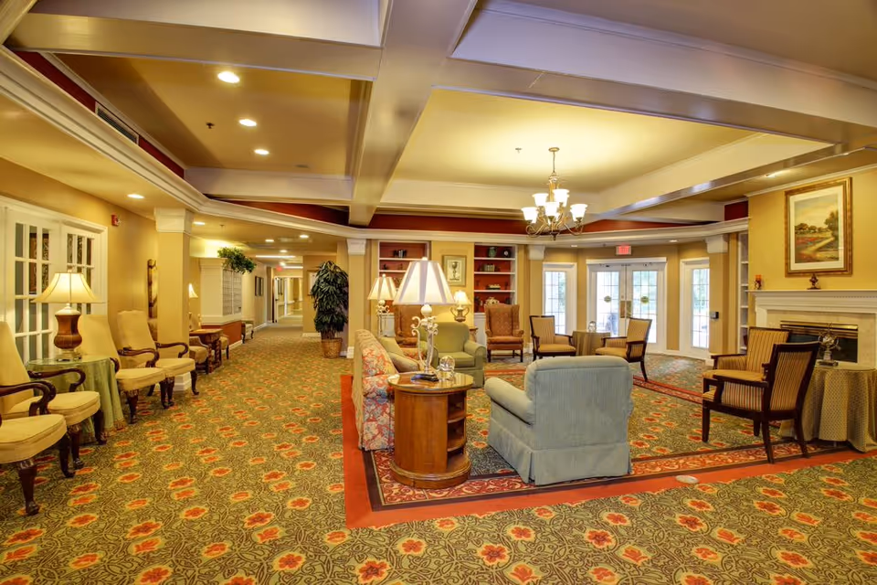 A spacious and warmly lit senior living facility common area with patterned carpet, multiple seating arrangements including armchairs and sofas, side tables with lamps, a chandelier, built-in shelves, and a fireplace. French doors lead to an outside area, and the hallway extends to the left.