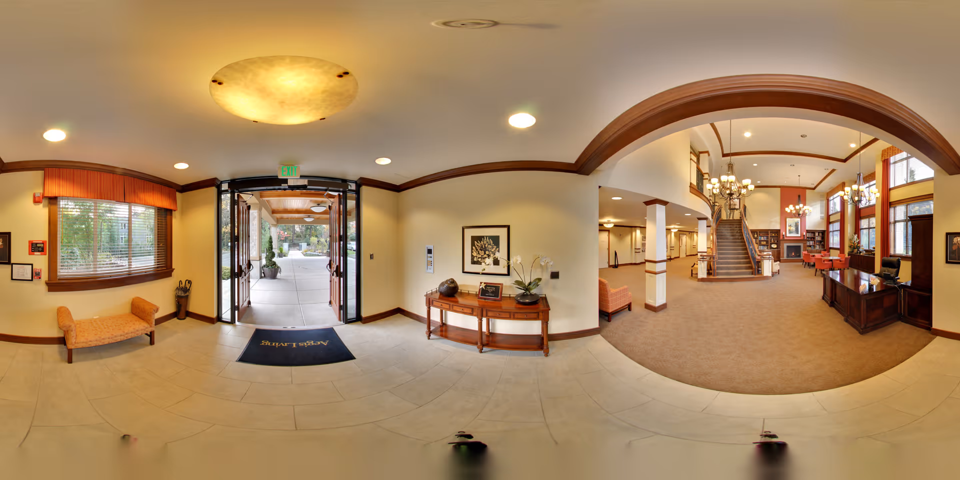 A spacious and well-lit senior living facility lobby with an open glass door leading outside. The interior features beige walls, wooden trim, a small wooden table with decorative items, a bench near a window with blinds, and a carpeted area with a staircase, chandelier lighting, and seating arrangements.