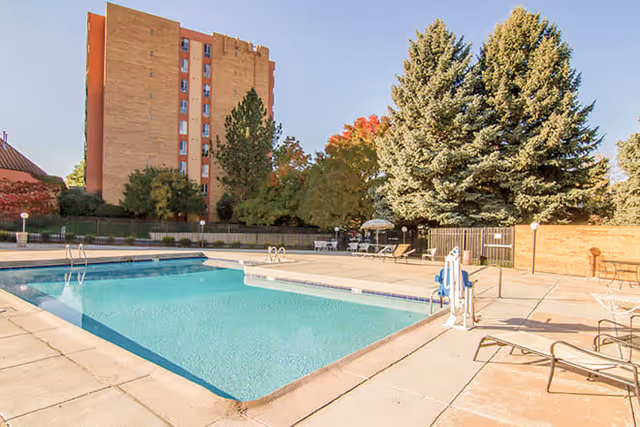 Outdoor swimming pool area with clear blue water, surrounded by a concrete deck with lounge chairs and tables. In the background, there is a tall brick building, trees with green and autumn-colored leaves, and a clear blue sky.