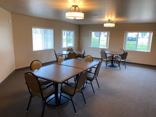 A small dining room with three tables, each surrounded by four chairs. The room has beige walls, carpeted floor, and three windows letting in natural light. Two ceiling lights are visible.