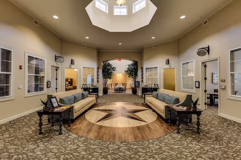 Spacious interior common area with two beige sofas facing each other on a patterned carpet. A large wooden star design is centered on the floor between the sofas. The room has beige walls, several windows, and doorways leading to other rooms. There is a skylight above, allowing natural light to brighten the space. Small tables with plants and picture frames are placed next to the sofas.