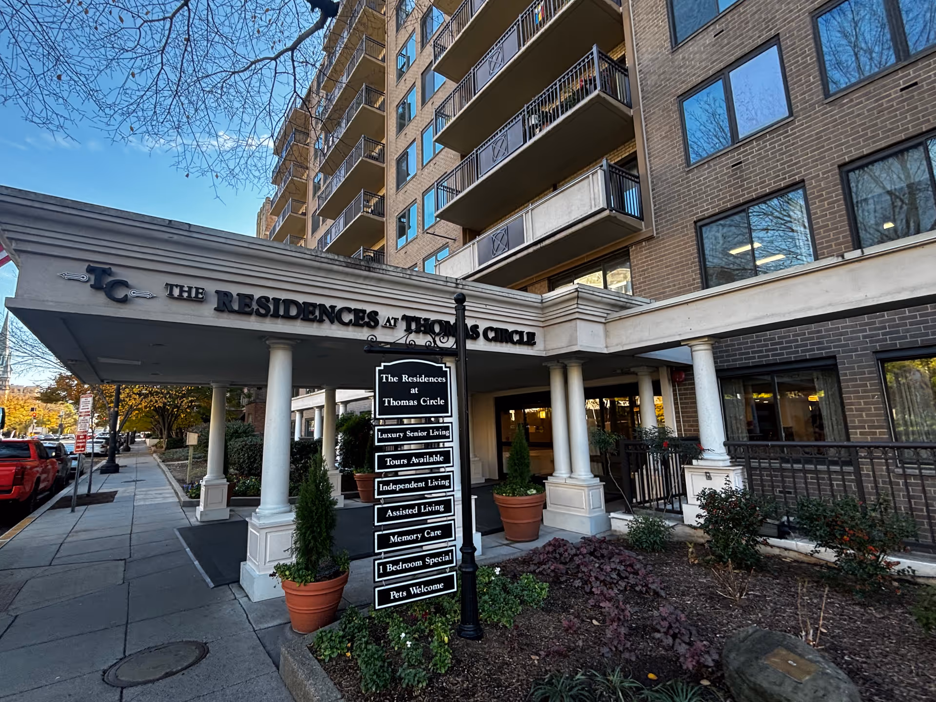 Exterior view of a multi-story senior living facility named The Residences at Thomas Circle, showing the entrance with white columns, a sign listing services such as luxury senior living, tours available, independent living, assisted living, memory care, 1 bedroom special, and pets welcome. The building has balconies and large windows, with some landscaping and a sidewalk in front.