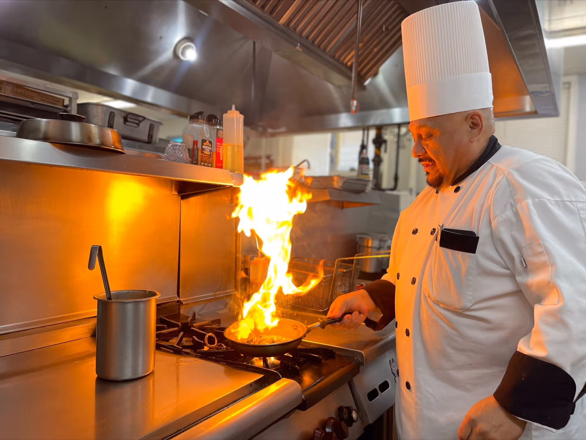 A chef in a white uniform flambés food in a pan over a flaming stovetop in a commercial kitchen.
