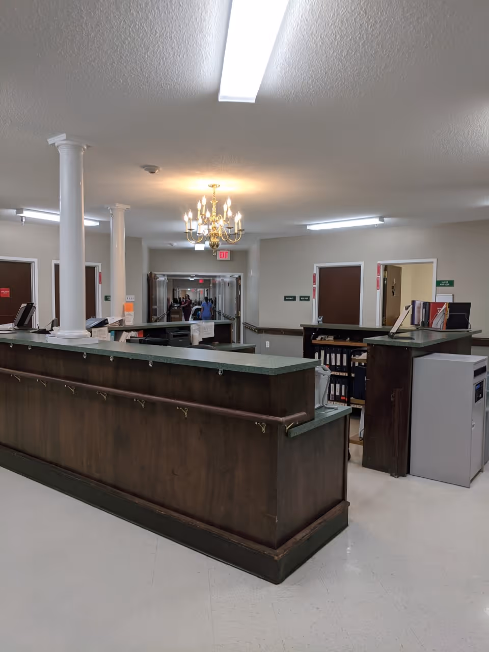 A reception/nurses' station with a long dark wood counter, columns and a chandelier in a senior facility hallway.