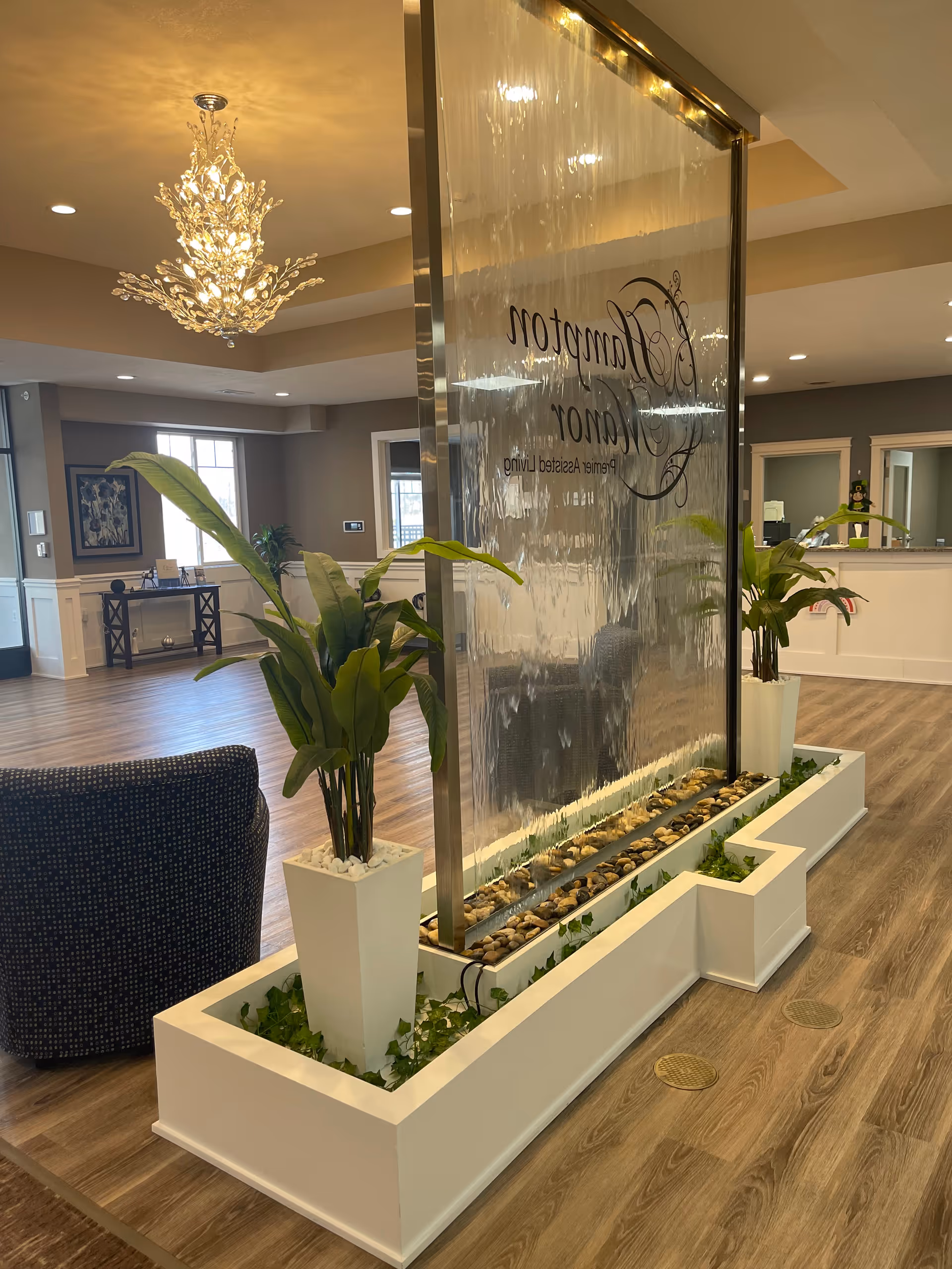 Interior view of a senior living facility with a decorative water wall feature in the center, flanked by two white planters with green plants. The room has wood flooring, a chandelier, recessed lighting, and a seating area with a dark upholstered chair. There is a reception desk visible in the background.