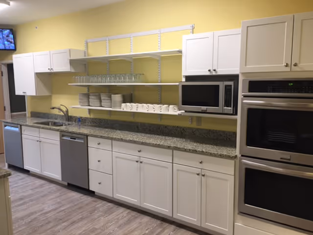 A clean and modern kitchen area with white cabinets, granite countertops, a stainless steel dishwasher, microwave, and double oven. Open shelves hold neatly stacked plates, cups, and glasses. The walls are painted yellow and the floor has wood-like vinyl flooring.
