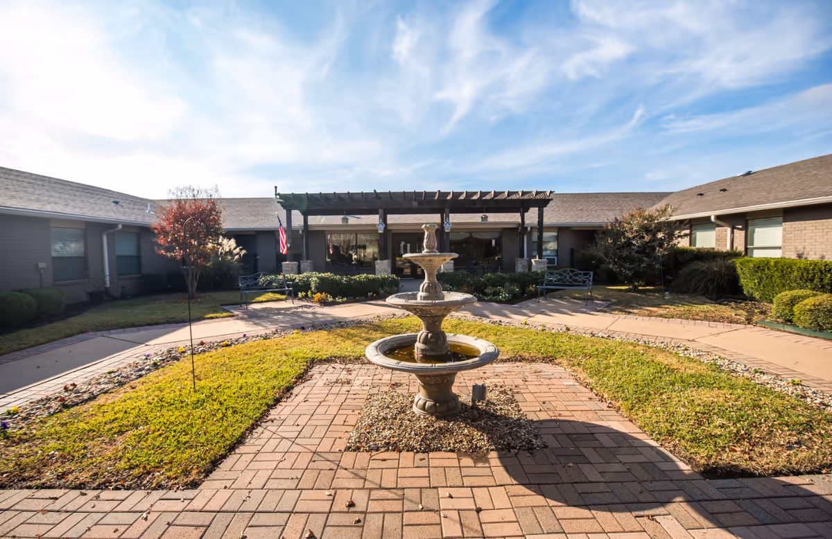 Courtyard with a three-tier fountain, brick walkway, pergola-covered seating area, and surrounding single-story building under a blue sky.