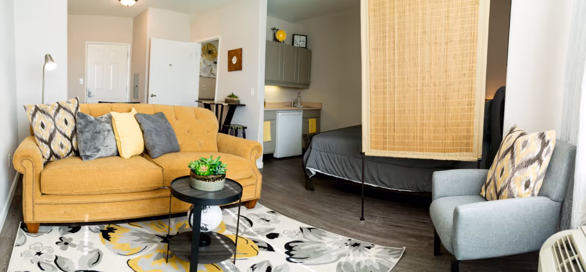 A cozy senior living space featuring a mustard yellow sofa with patterned and solid pillows, a small round black coffee table with a potted succulent, a gray armchair with a patterned pillow, and a floral area rug. In the background, there is a small kitchenette with a mini fridge and cabinets, a bed partially hidden by a bamboo room divider, and a white door.