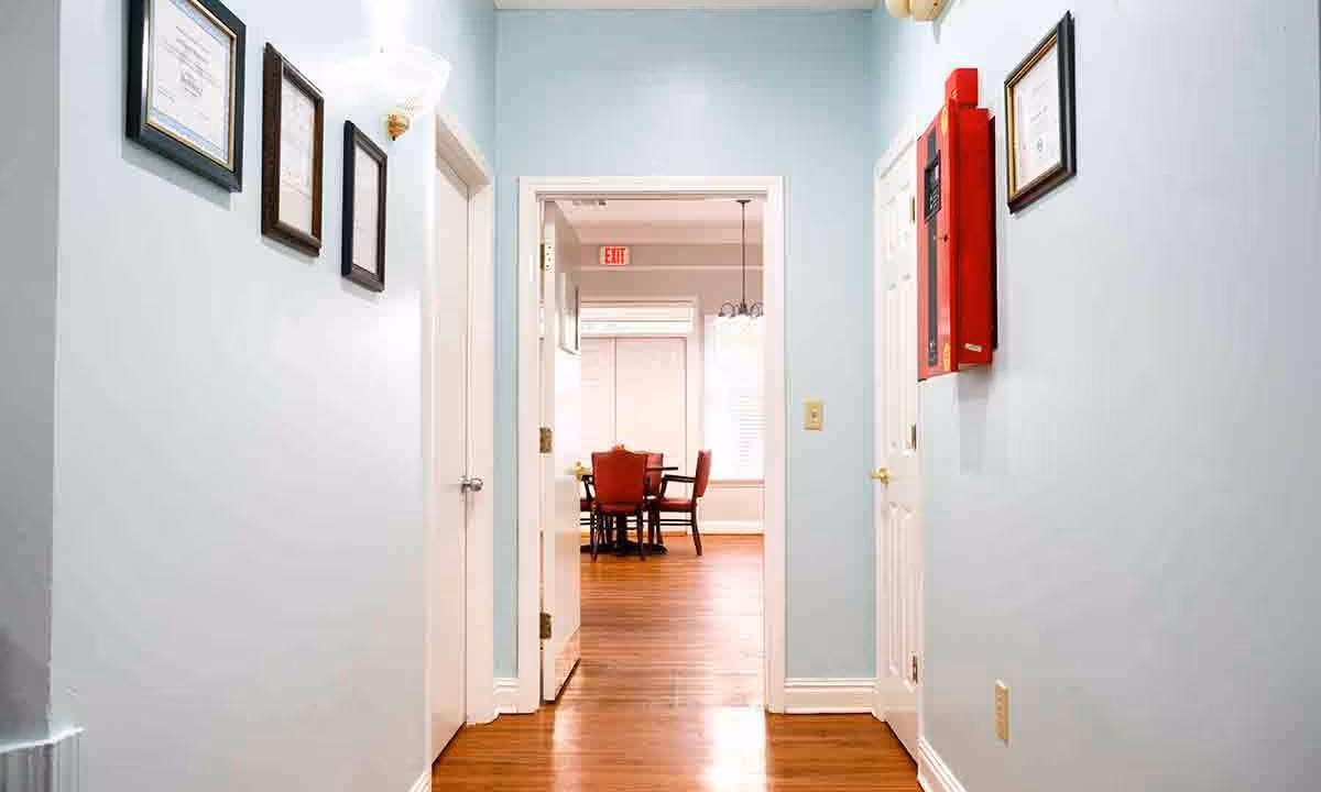 View down a hallway with light blue walls and wooden floor leading to a dining area with a table and red chairs. The hallway has framed certificates on the walls and a red fire safety box on the right side.