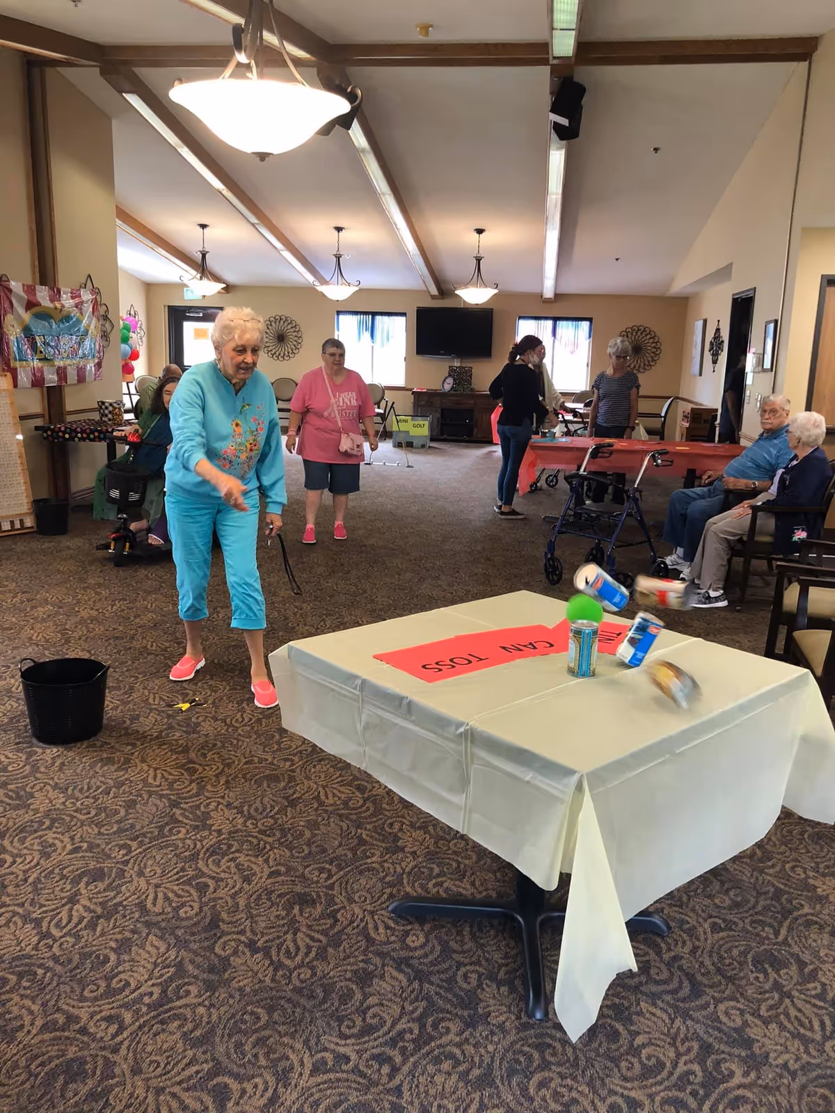 A group of elderly people and a few caregivers are in a spacious common room with carpeted floor and ceiling lights. An elderly woman in a turquoise outfit is tossing bean bags towards a table with a sign that reads 'CAN TOSS'. Other residents and staff are watching or sitting nearby, with some seated and some standing. The room has windows, chairs, and a television mounted on the far wall.