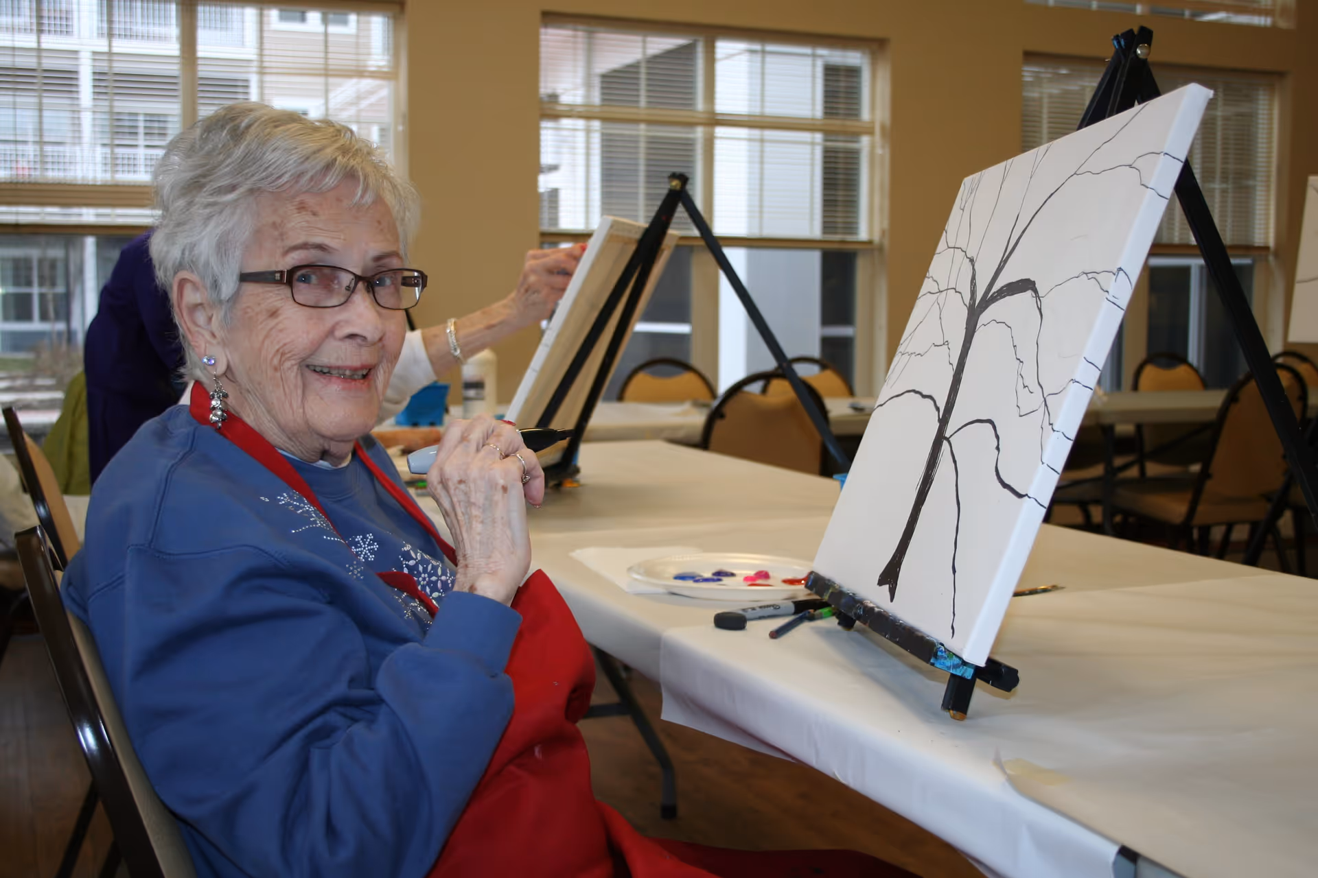 An elderly woman wearing glasses and a red apron is sitting at a table in a well-lit room, smiling at the camera while painting a tree on a canvas. The table has painting supplies including a palette with paint and brushes. Other people are also painting in the background.