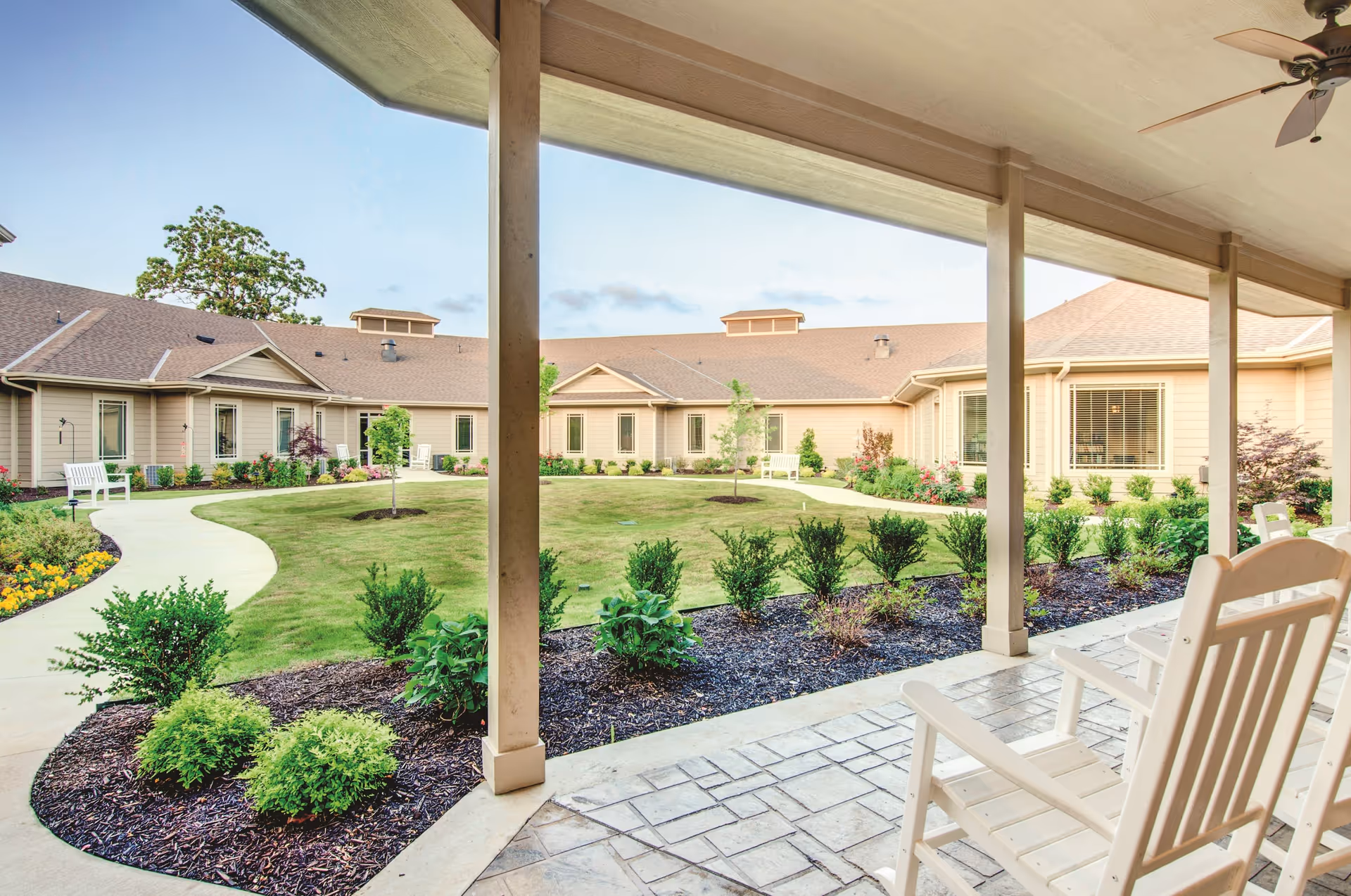 View from a covered patio with white rocking chairs overlooking a landscaped courtyard with green grass, small trees, shrubs, and a curved walkway surrounded by a single-story beige building under a clear sky.