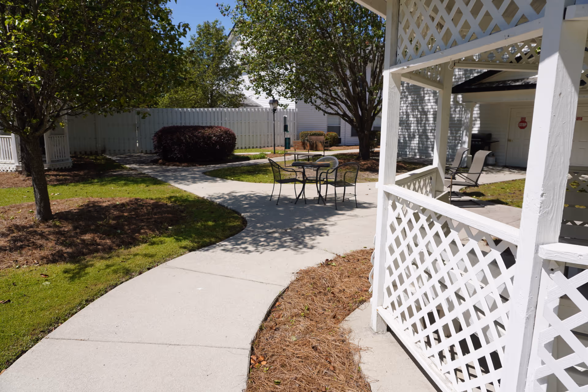 Outdoor area with a curved concrete pathway, green grass, trees, and bushes. There is a white lattice gazebo on the right side and a small round table with four chairs in the center. A white fence and a building are visible in the background under a clear blue sky.
