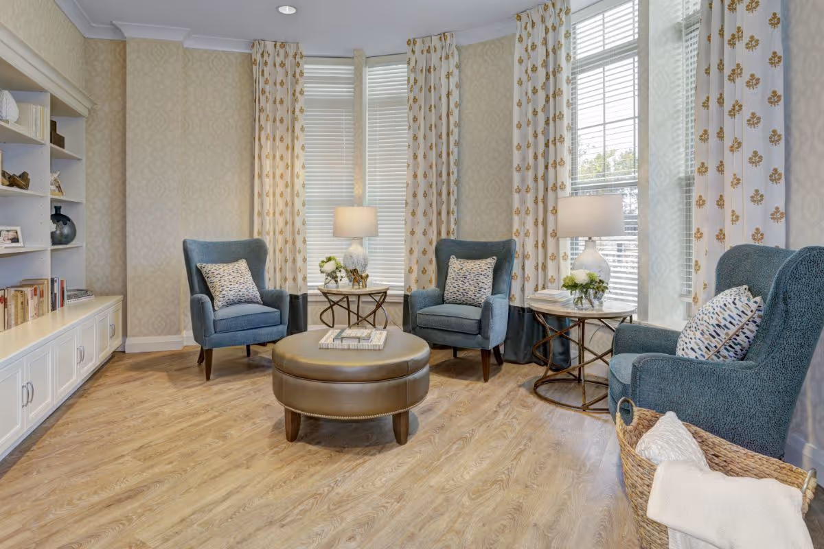 A cozy living room area with three blue upholstered armchairs, each with a patterned cushion. Two round side tables with white lamps and floral arrangements are placed near the windows, which have white blinds and curtains with a gold floral pattern. A round brown leather ottoman sits in the center on a wood floor. A built-in white bookshelf with books and decorative items is on the left wall, and a woven basket with a white blanket is on the floor near one chair.