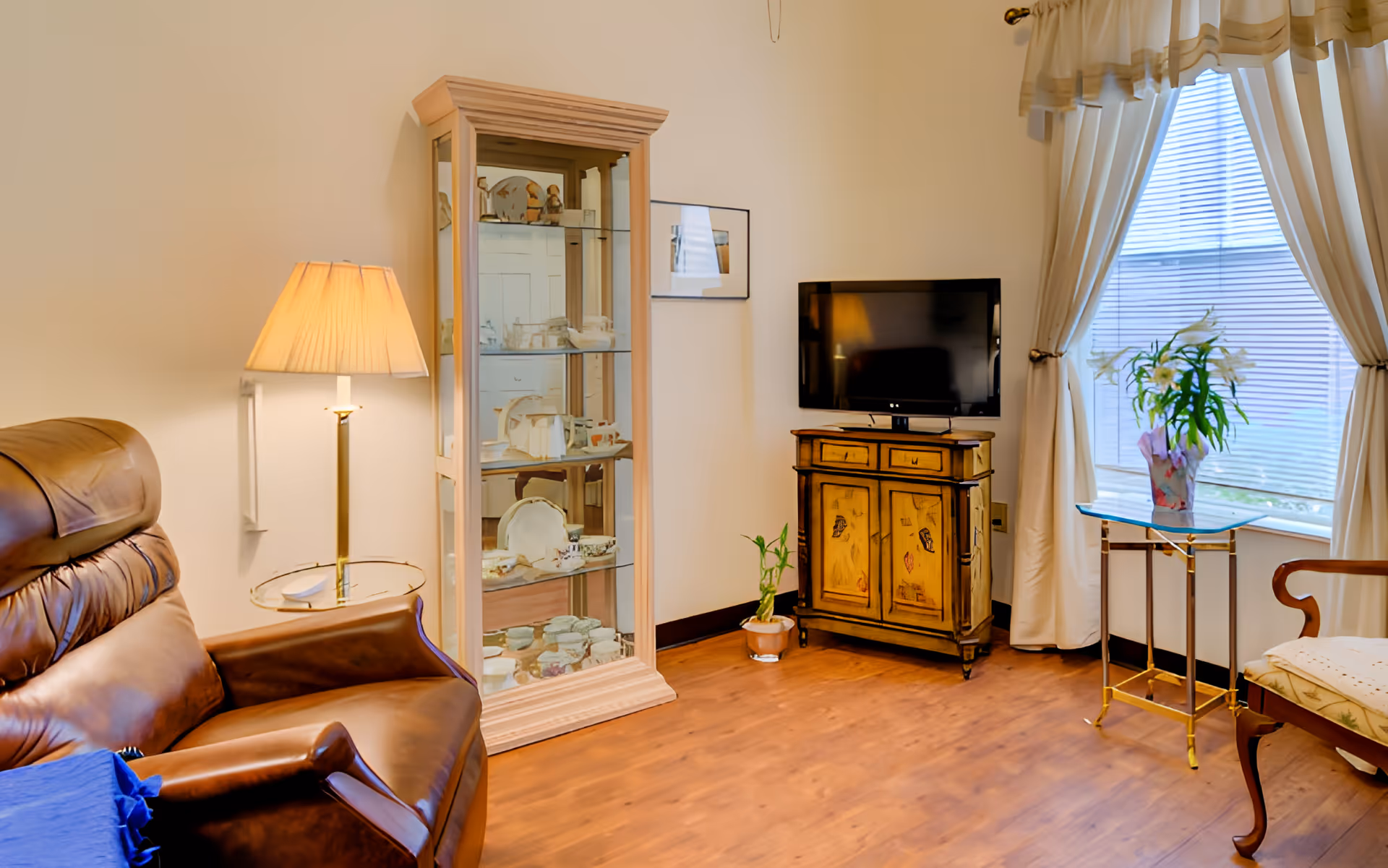 A cozy living room with a brown leather recliner chair, a floor lamp with a beige shade, a glass display cabinet filled with decorative items, a small wooden cabinet with a flat-screen TV on top, a potted plant on the floor, a glass side table with a vase of flowers, and a window with white curtains allowing natural light to enter.