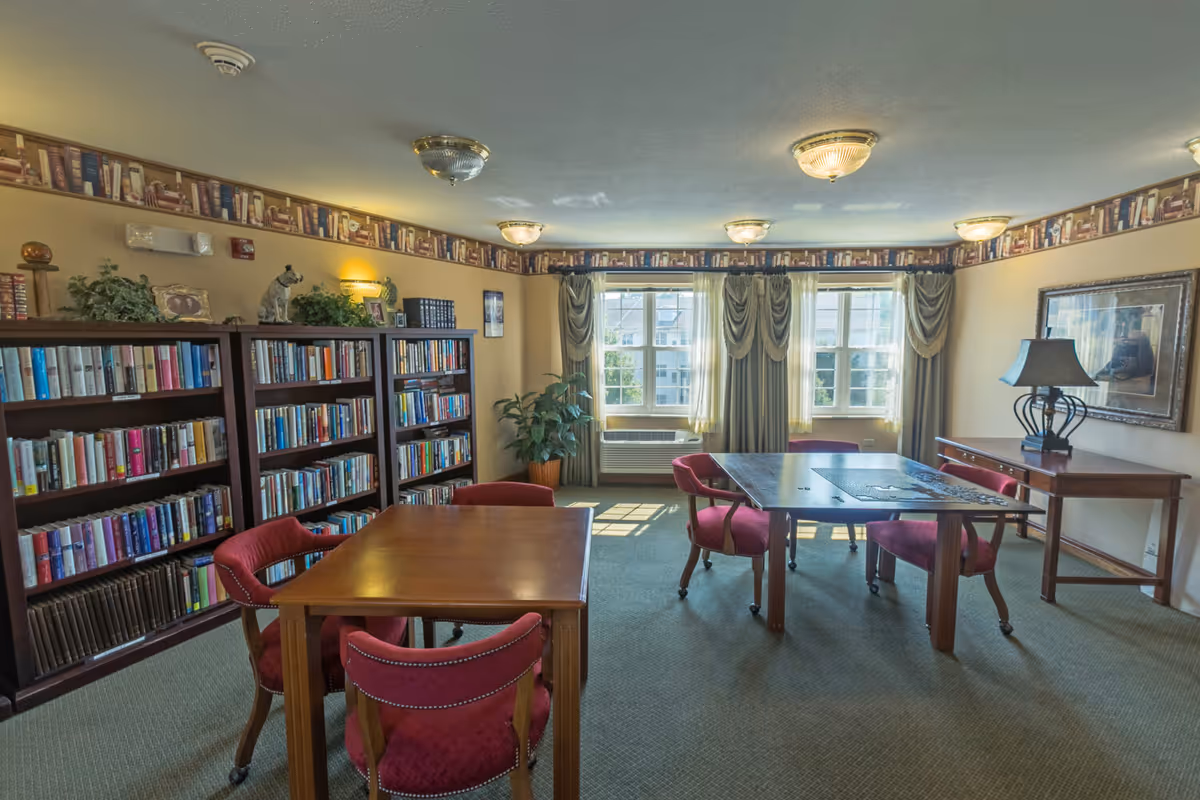 A cozy reading or activity room with bookshelves filled with books along one wall, two wooden tables with red upholstered chairs around them, a large window with draped curtains letting in natural light, a side table with a lamp, and decorative plants and framed pictures on the walls.