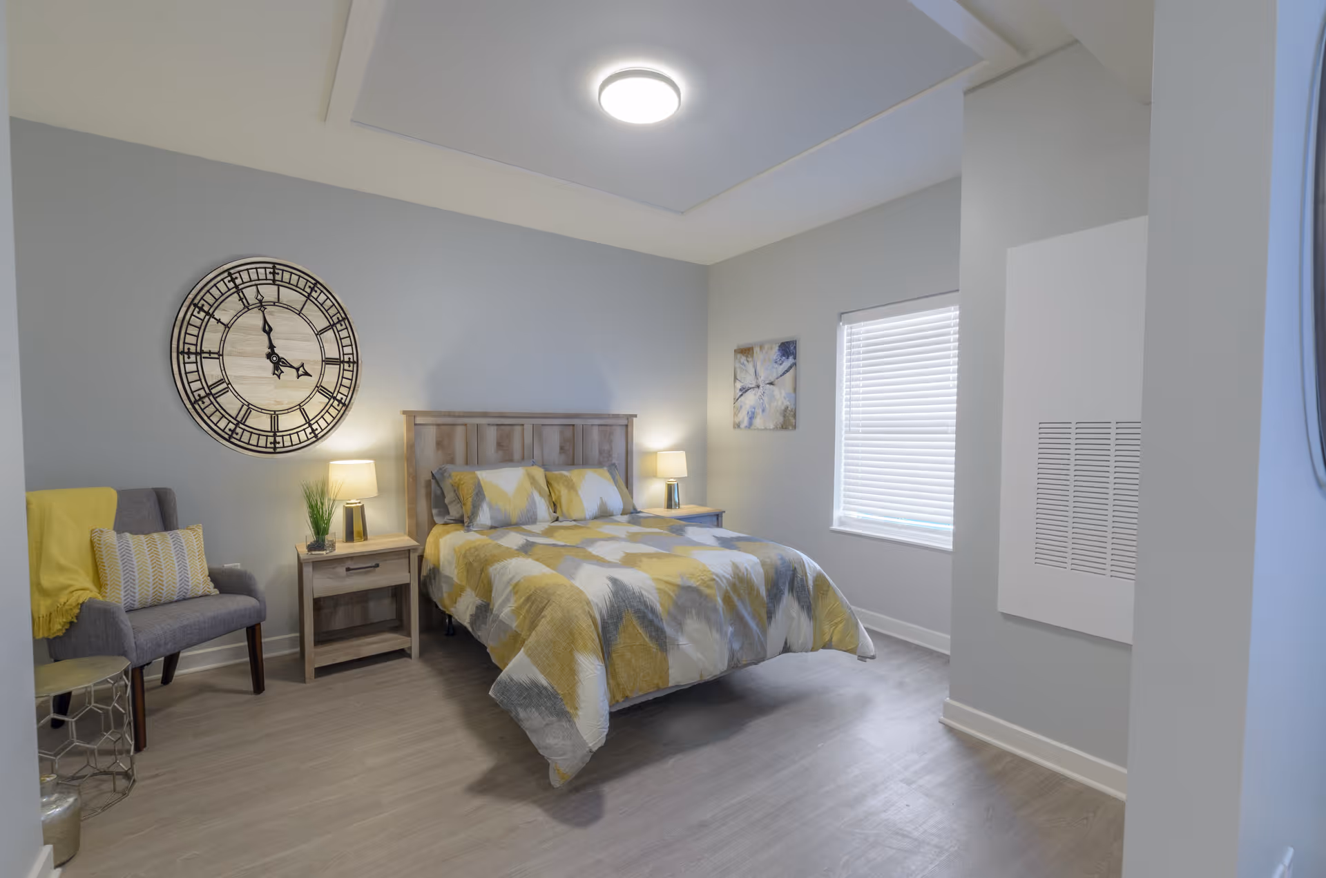 Bright, modern bedroom featuring a wooden bed with yellow-gray bedding, two nightstands with lamps, an accent chair, and a large wall clock.