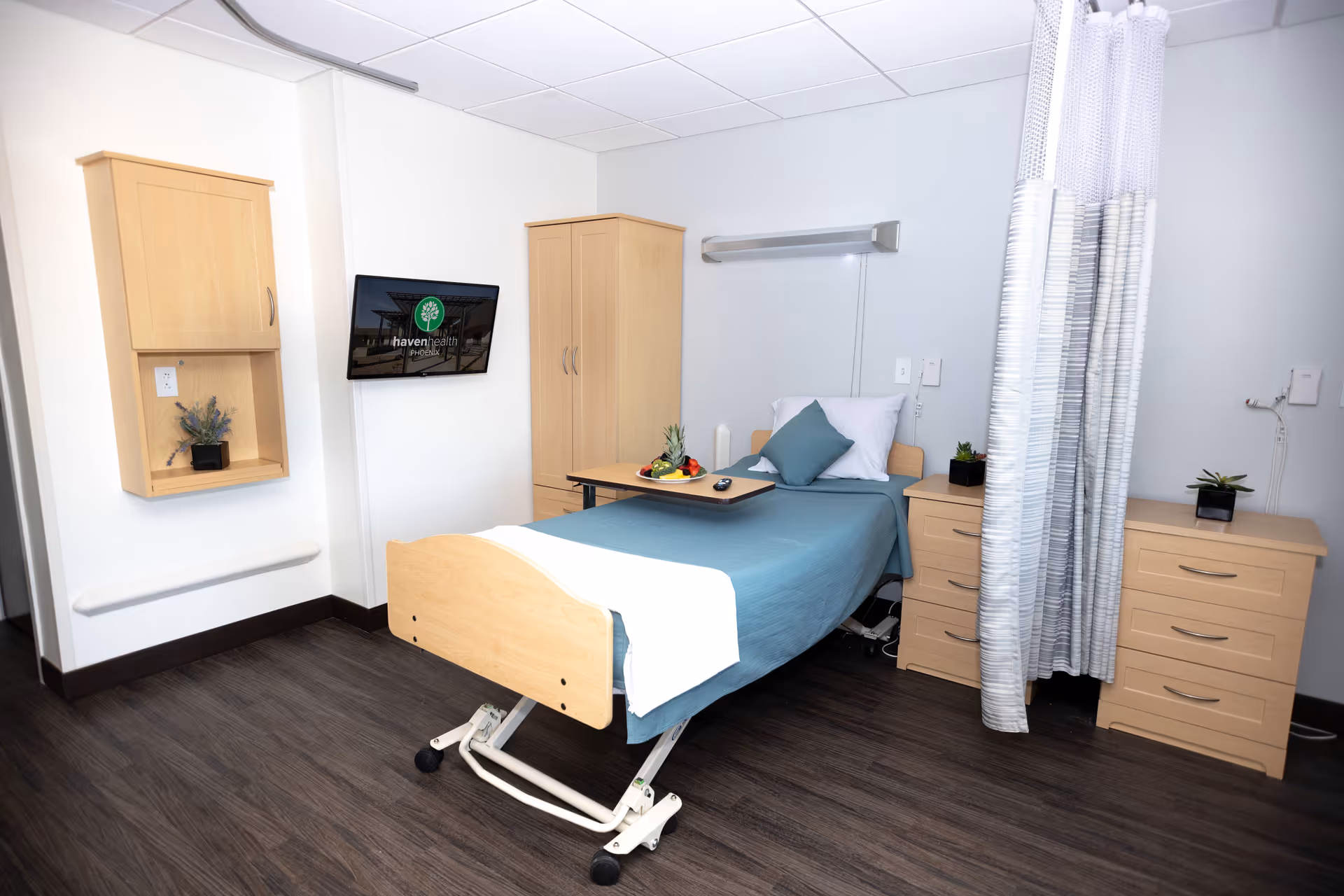 A clean and bright patient room in a senior living facility with a single hospital-style bed covered with a teal blanket and white pillow. The room features light wood furniture including a wardrobe, two dressers, and a wall-mounted cabinet. A tray with fruit and a remote control is placed on the bed. A flat-screen TV mounted on the wall displays the Haven Health Phoenix logo. A privacy curtain is partially drawn to the side.