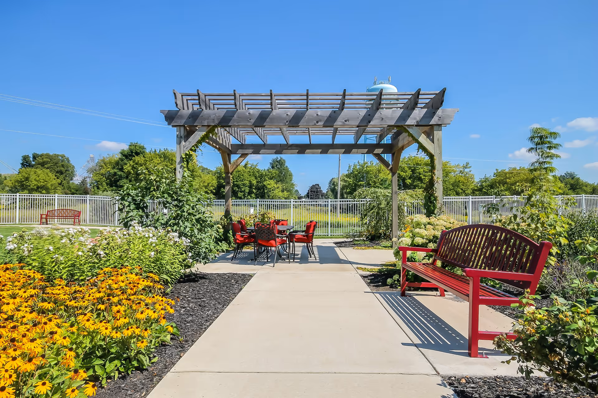 Outdoor garden area at Lakeview Terrace Assisted Living featuring a wooden pergola with a table and red chairs underneath, surrounded by lush greenery, colorful flowers, and a red bench along a paved walkway under a clear blue sky.