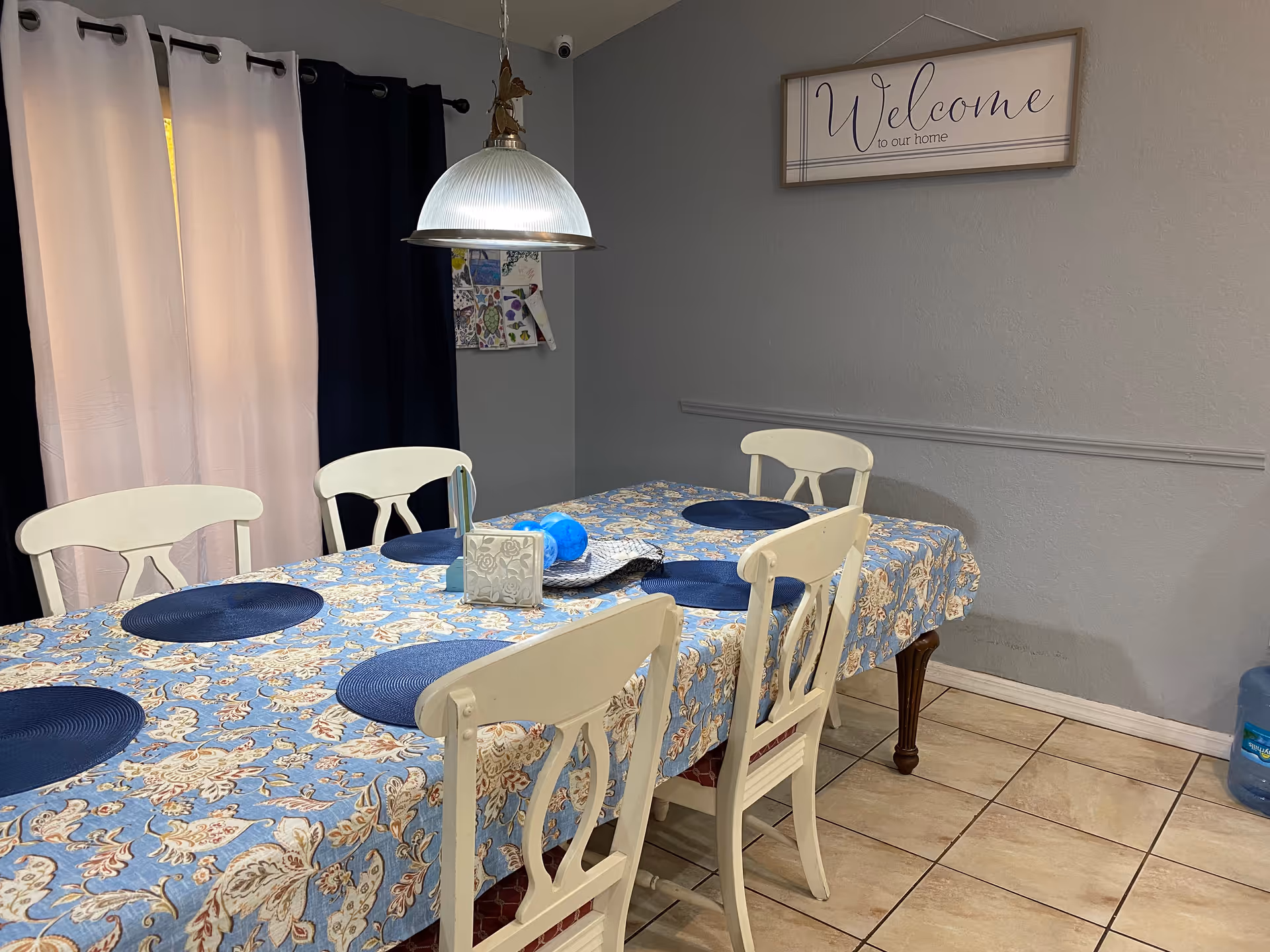 A dining room with a rectangular table covered with a blue floral tablecloth and six white chairs around it. The table has blue placemats, a tissue box, and decorative items. Behind the table are two-tone curtains in light pink and dark blue. A hanging light fixture is above the table, and a wall sign reads 'Welcome to our home'. The floor is tiled, and a water dispenser is visible in the corner.