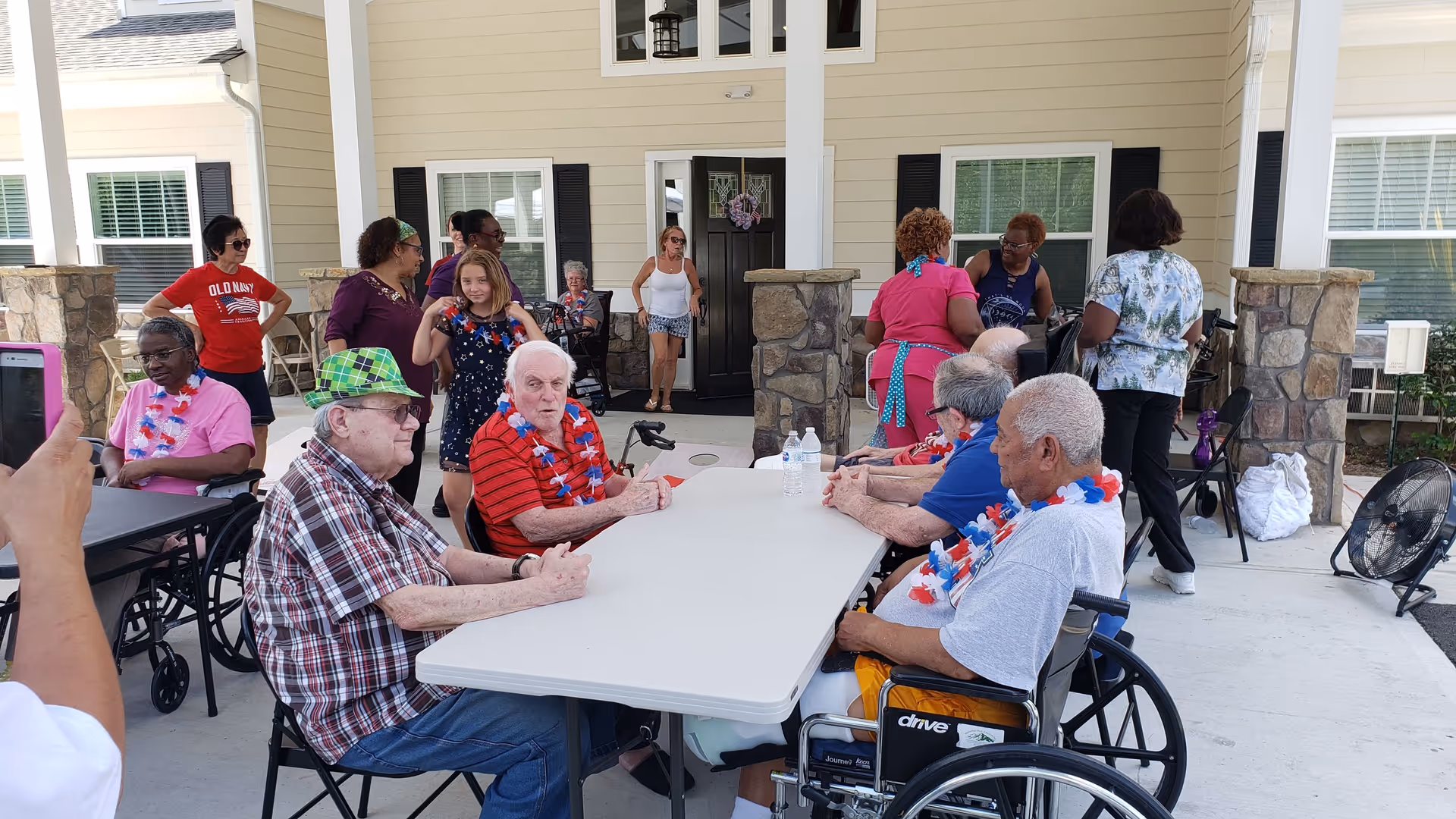 A group of elderly people, some in wheelchairs, sitting around a white table outside a senior care facility. Several staff members and visitors are standing and interacting in the background near the building entrance. The elderly individuals are wearing colorful leis and appear to be enjoying a social gathering.