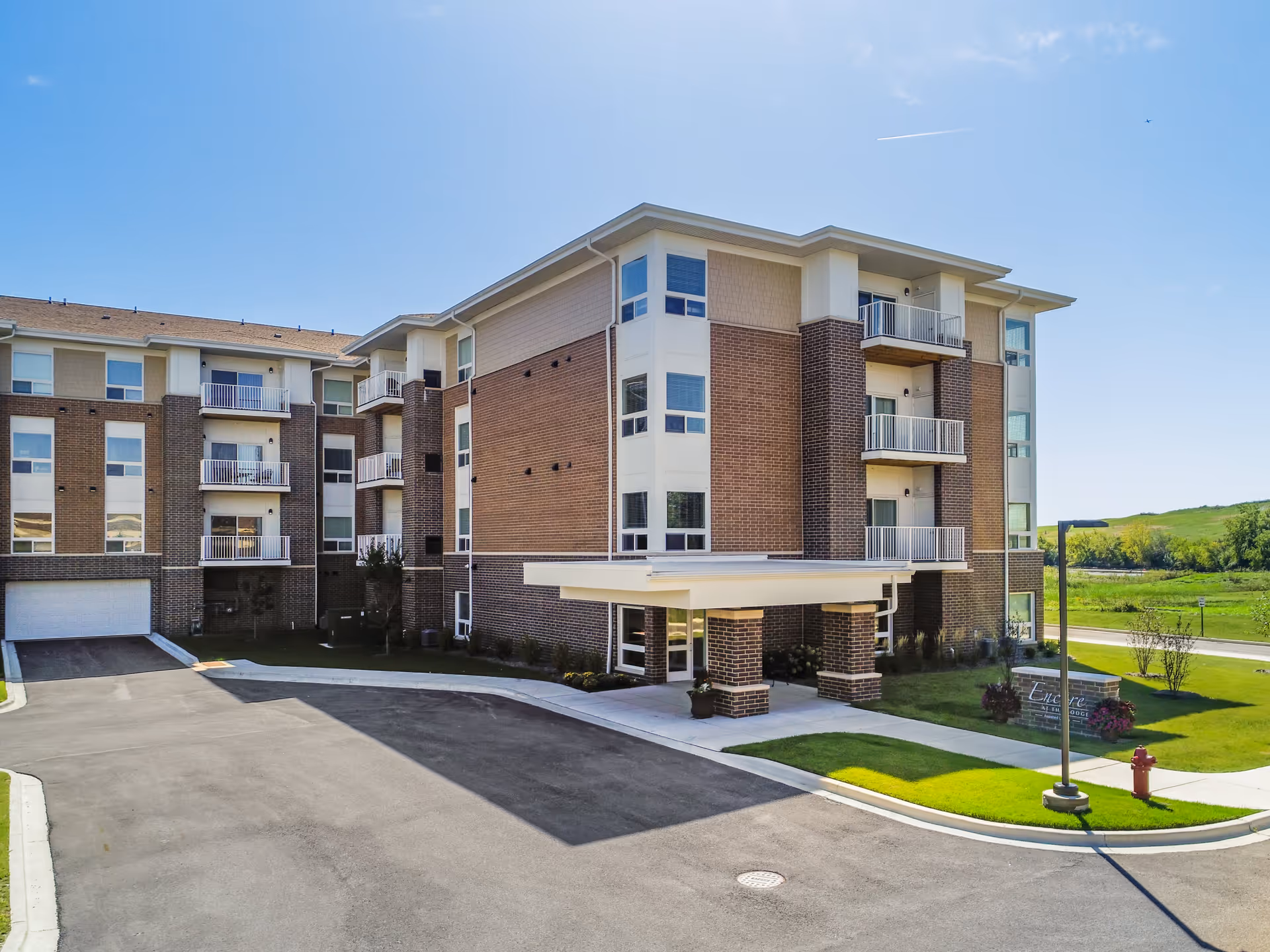 Exterior view of a modern four-story senior living facility building named Encore at The Lodge, featuring brick and beige siding, balconies, a covered entrance, a driveway, and well-maintained green landscaping under a clear blue sky.