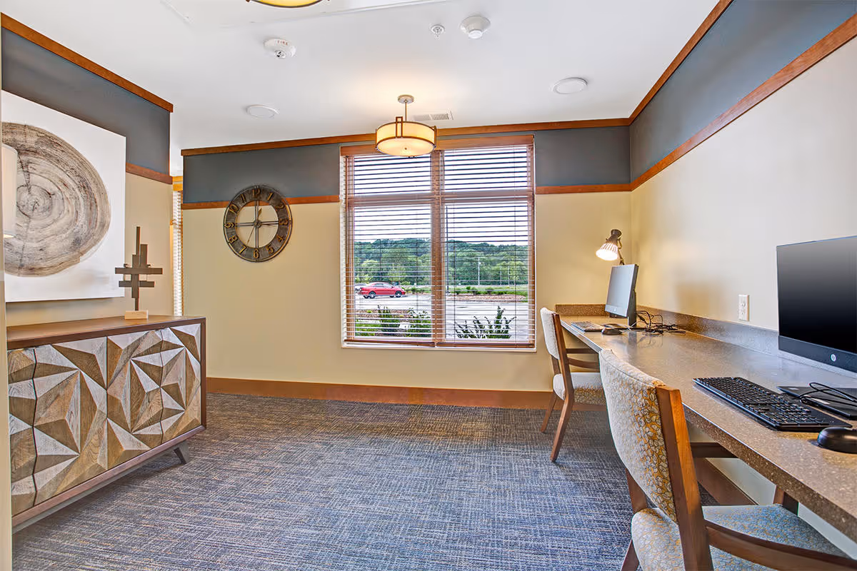 A well-lit interior room with a large window featuring wooden blinds overlooking a parking lot and greenery. The room has a patterned carpet, a decorative wooden cabinet with geometric designs, a wall clock, and a long counter with two chairs and desktop computers. The walls are painted in two tones with a wooden trim.