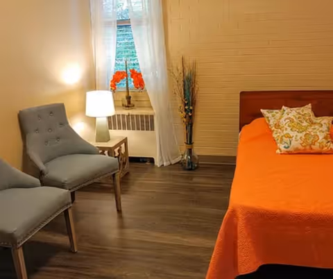 A cozy bedroom with a bed covered in an orange blanket and two decorative pillows. Two gray upholstered chairs are placed near a small wooden side table with a lamp. A window with sheer white curtains and a vase with orange flowers is visible, along with a tall vase with decorative branches in the corner.