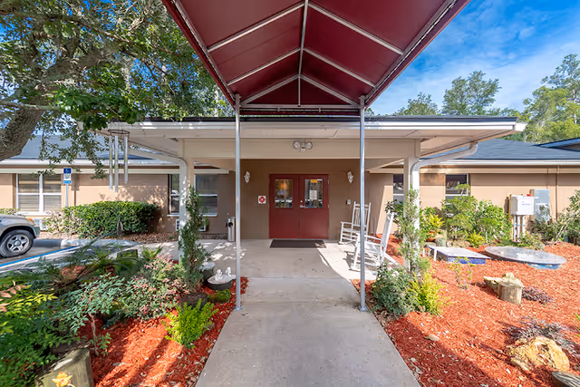 Covered walkway with a red canopy leading to double red entrance doors of a single-story senior living facility with landscaped beds and rocking chairs.
