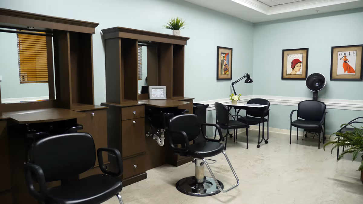 Interior of a salon area with two black salon chairs in front of wooden styling stations with mirrors. There is a small table with two black chairs, a desk lamp, and three framed Vogue magazine covers on the wall. A hair dryer chair and a green plant are also visible.