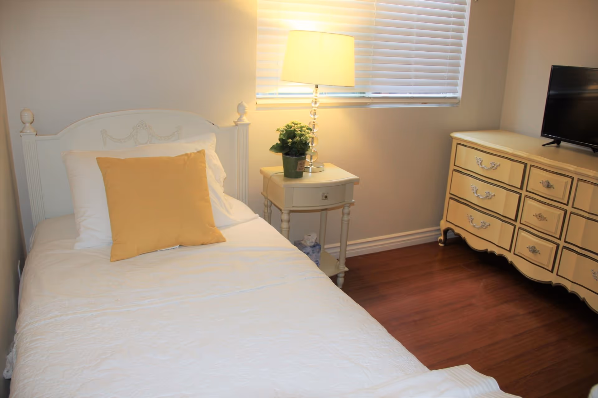 A cozy bedroom with a single white bed featuring a yellow pillow and white bedding. Next to the bed is a small white nightstand with a potted plant and a lit lamp. A window with closed blinds is above the nightstand. On the right side of the room is a cream-colored dresser with a flat-screen TV on top. The floor is wooden.