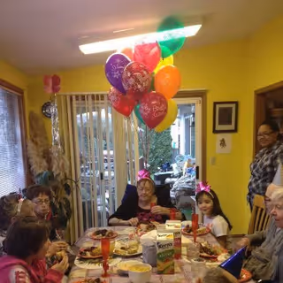 A group of people gathered around a dining table decorated with a tablecloth, party hats, and colorful balloons. The table has plates of food, snacks, and drinks. The setting appears to be indoors with large windows and sliding glass doors in the background. A woman stands to the right, smiling.