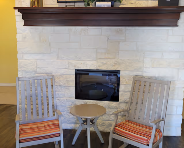 Two wooden chairs with striped cushions positioned on either side of a small round table in front of a stone fireplace with a dark wooden mantel.