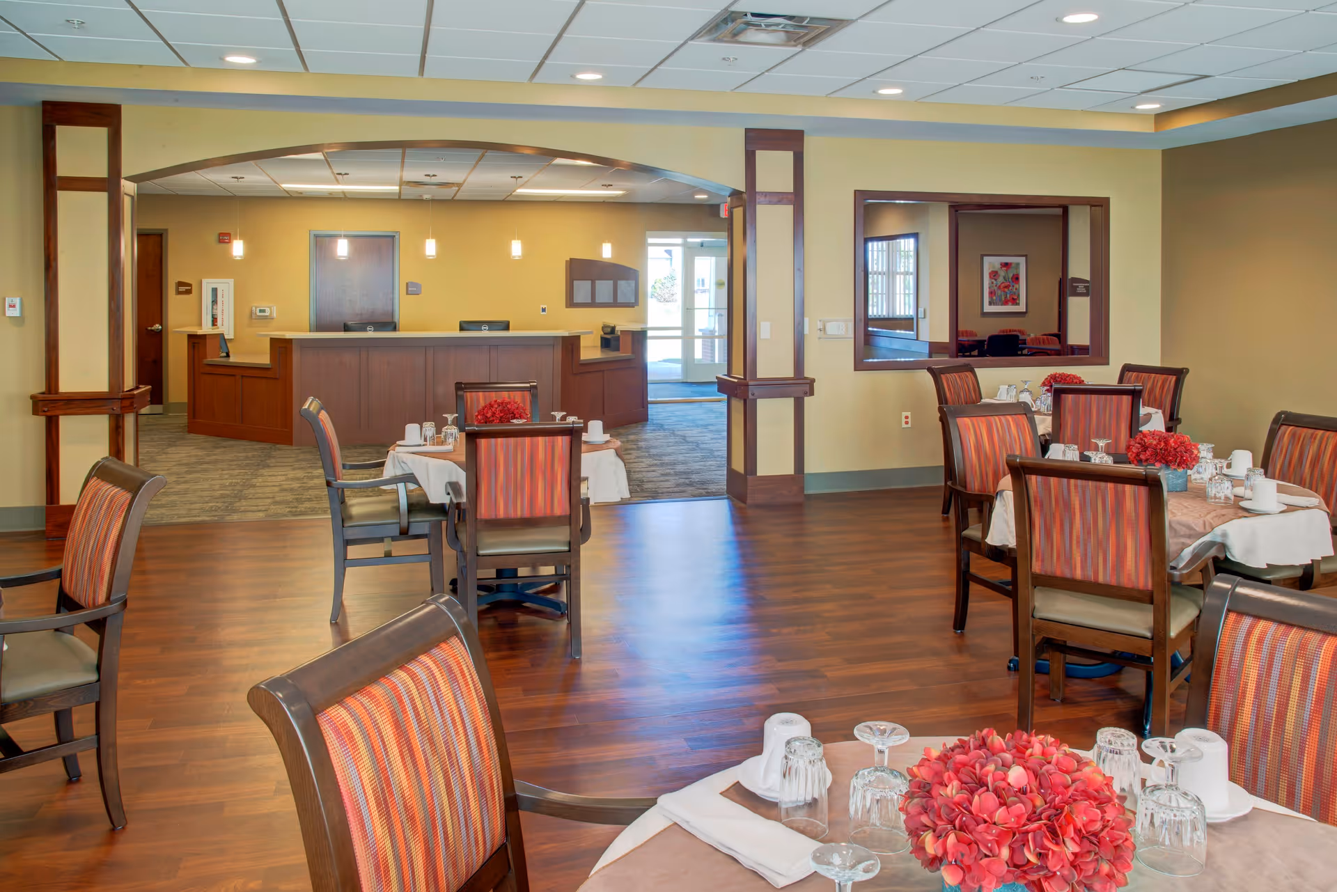 A dining area in a senior living facility with several tables set with white tablecloths, glassware, and red floral centerpieces. The room has wooden floors and chairs with striped upholstery. In the background, there is a reception desk with two chairs and a doorway leading outside.