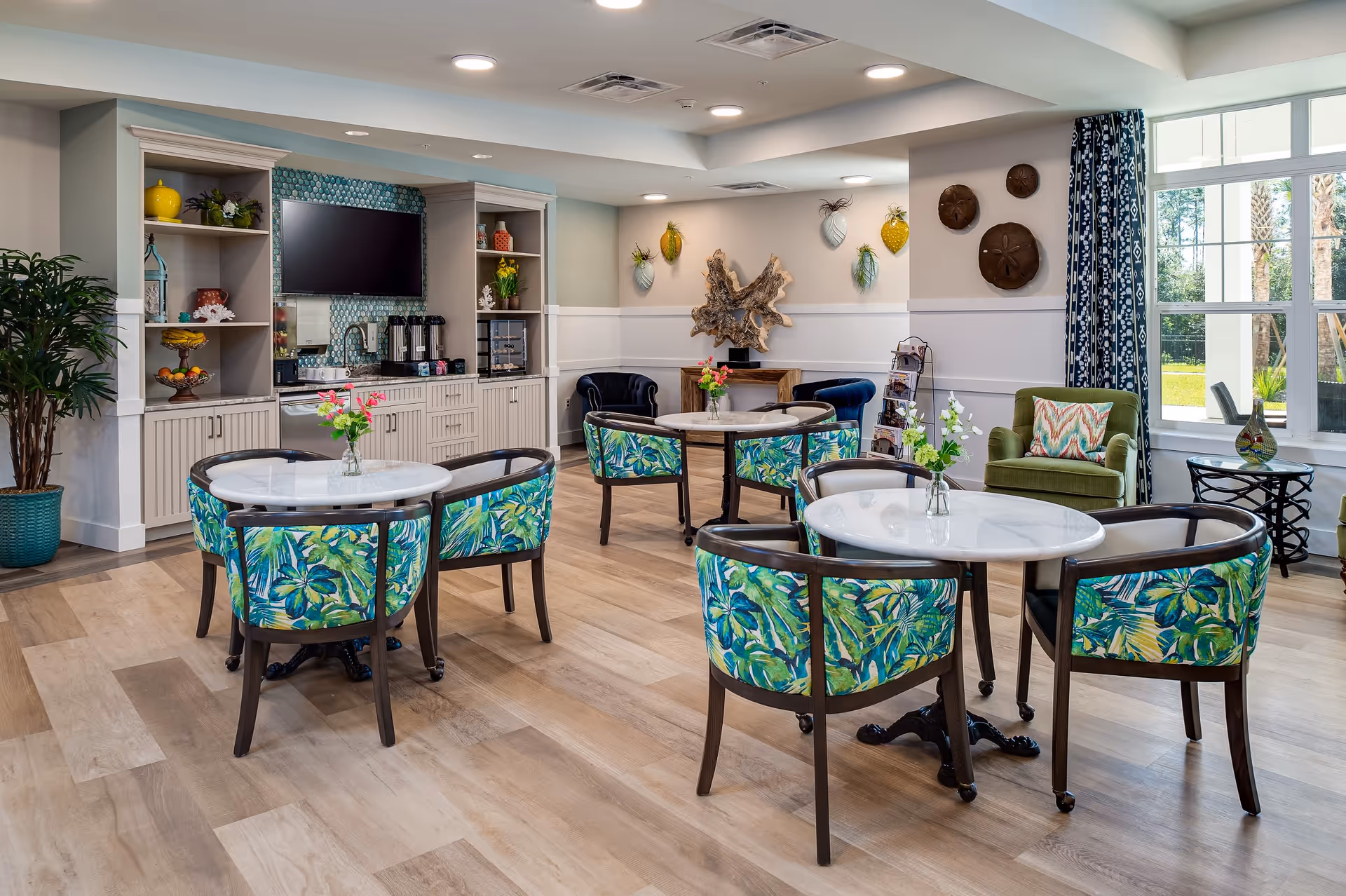 A bright and inviting common area with several round tables surrounded by chairs upholstered in a green and blue leaf pattern. The room features a built-in cabinet with a coffee station and a mounted flat-screen TV. Decorative wall art and plants add a cozy touch, and large windows let in natural light with a view of greenery outside.