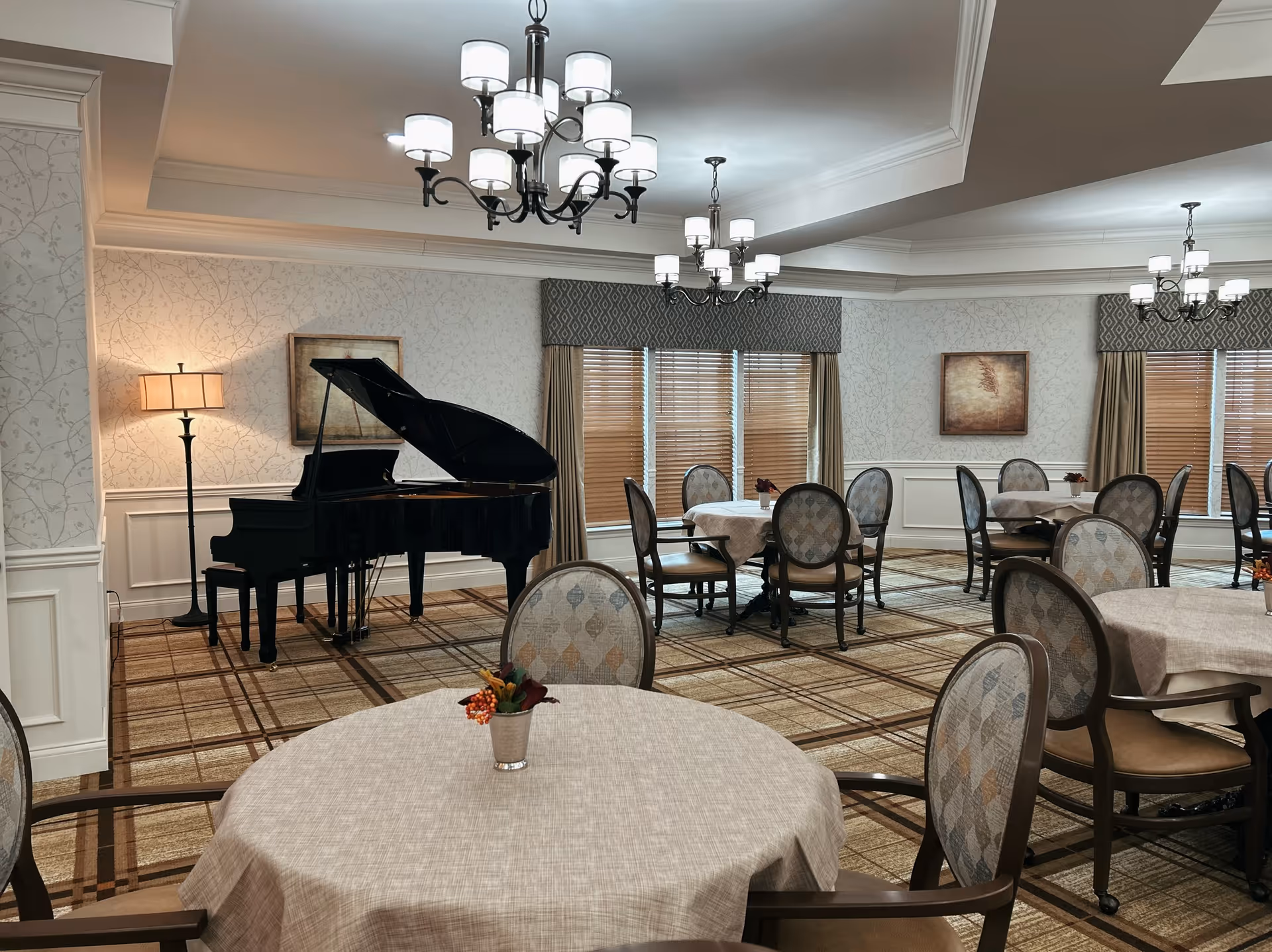 A dining room with multiple round tables covered with beige tablecloths and surrounded by cushioned chairs. A black grand piano is positioned near the wall with a floor lamp beside it. The room has patterned carpet, wallpaper with a subtle design, and windows with wooden blinds and valances. Several chandeliers hang from the ceiling providing light.