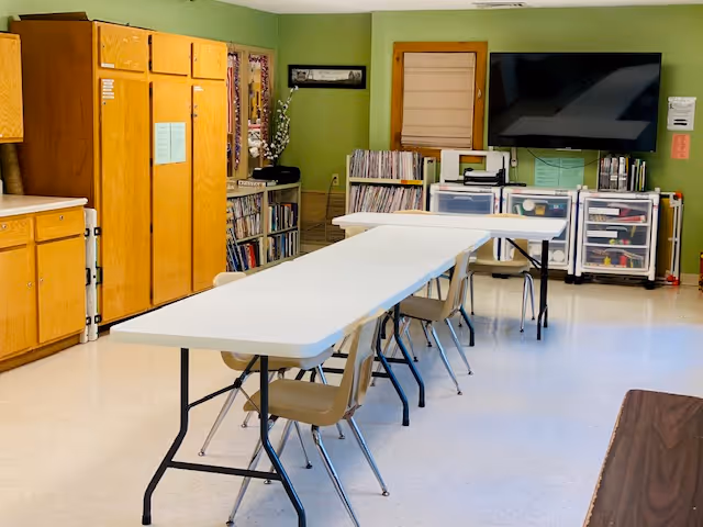 A well-lit room with green walls featuring two long white folding tables arranged in an L shape with beige chairs around them. On the left side, there are wooden cabinets and shelves filled with books and materials. On the far wall, there is a large flat-screen TV mounted above storage units containing various supplies. The floor is light-colored and shiny.