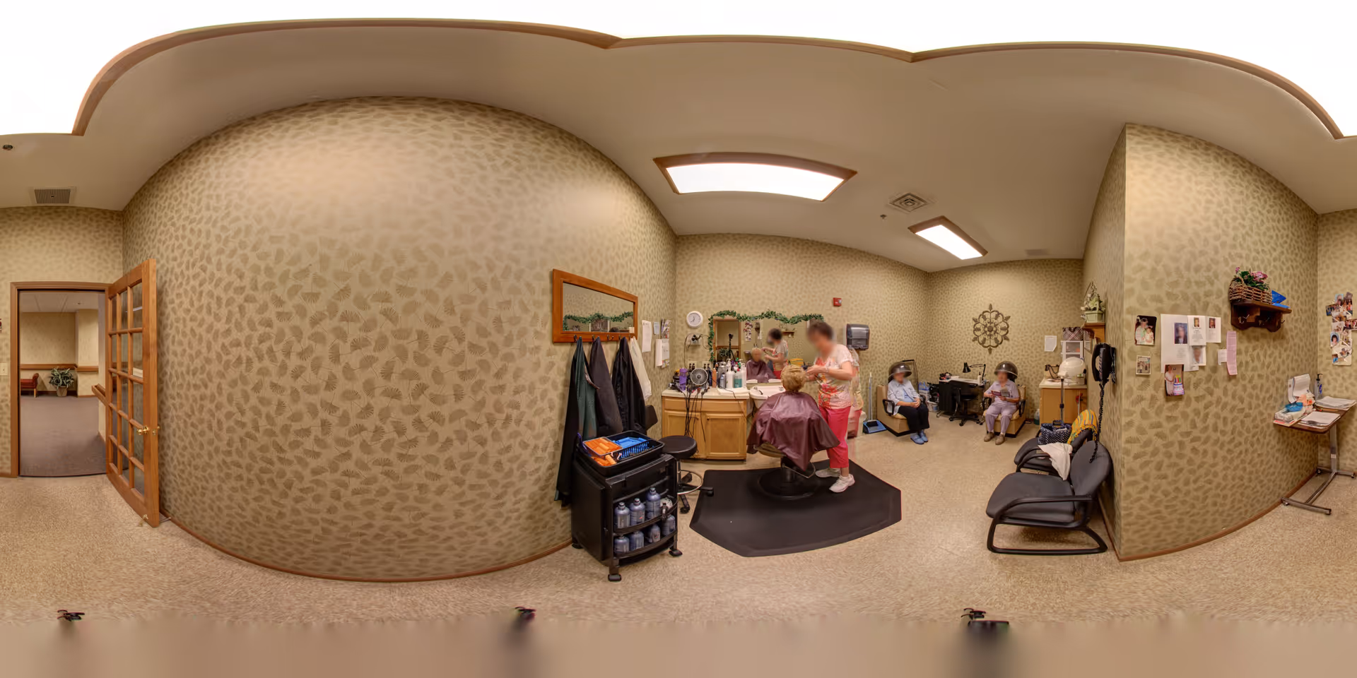 A salon room inside a senior living facility with a hairdresser cutting a resident's hair. The room has beige patterned wallpaper, a mirror, salon chairs, and various hair care products. Two other residents are seated in the background, waiting. There are coats hanging on the wall and a small table with magazines and decorations.