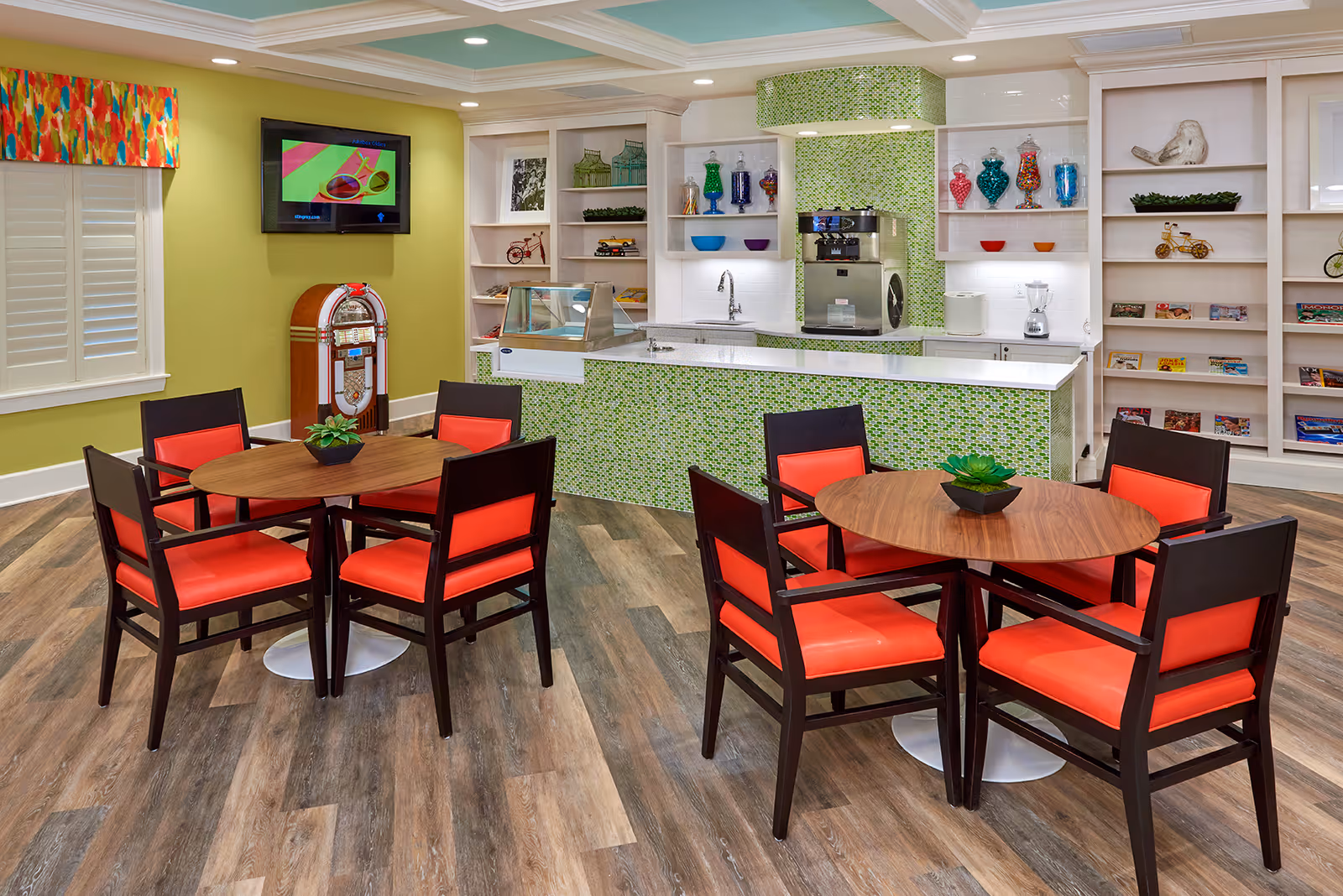 Community dining area with round wooden tables and red-upholstered chairs in front of a small kitchenette with green mosaic backsplash and shelving.
