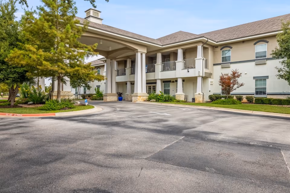 Front exterior of a two-story senior living building with a covered porte-cochere, balconies, landscaped trees, and a paved driveway.