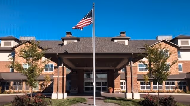 Front exterior view of Deerfield Springs Retirement Resort building with a covered entrance, brick facade, and an American flag on a flagpole in front under a clear blue sky.
