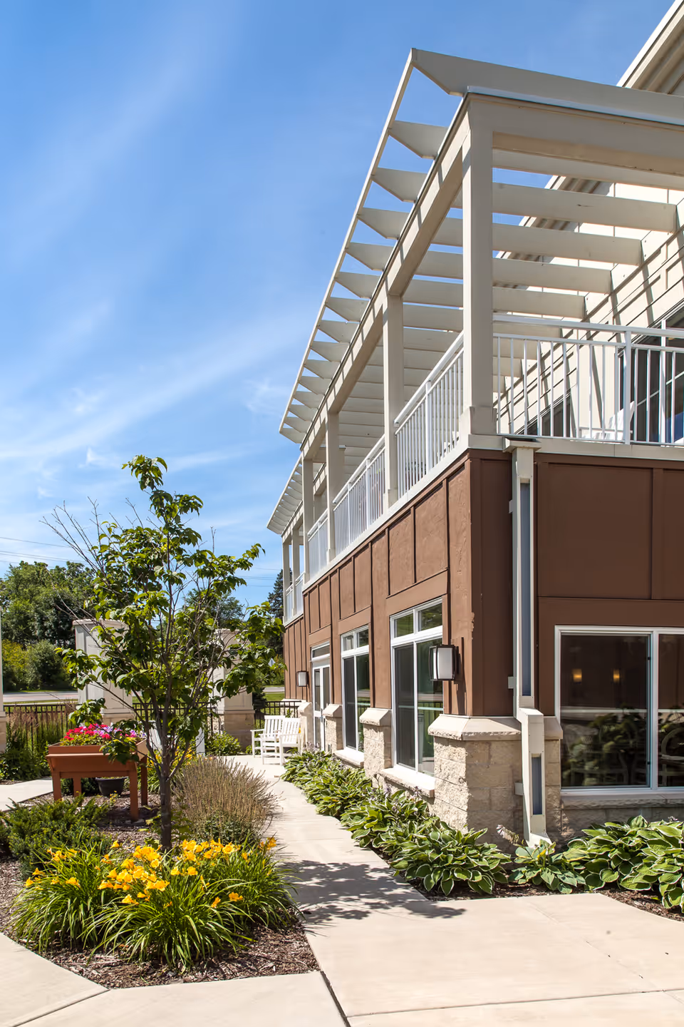 Outdoor view of a senior living facility building with a brown exterior and white balcony railings under a clear blue sky. There is a paved walkway surrounded by green plants and yellow flowers, with a small tree and a planter box with pink flowers along the path.