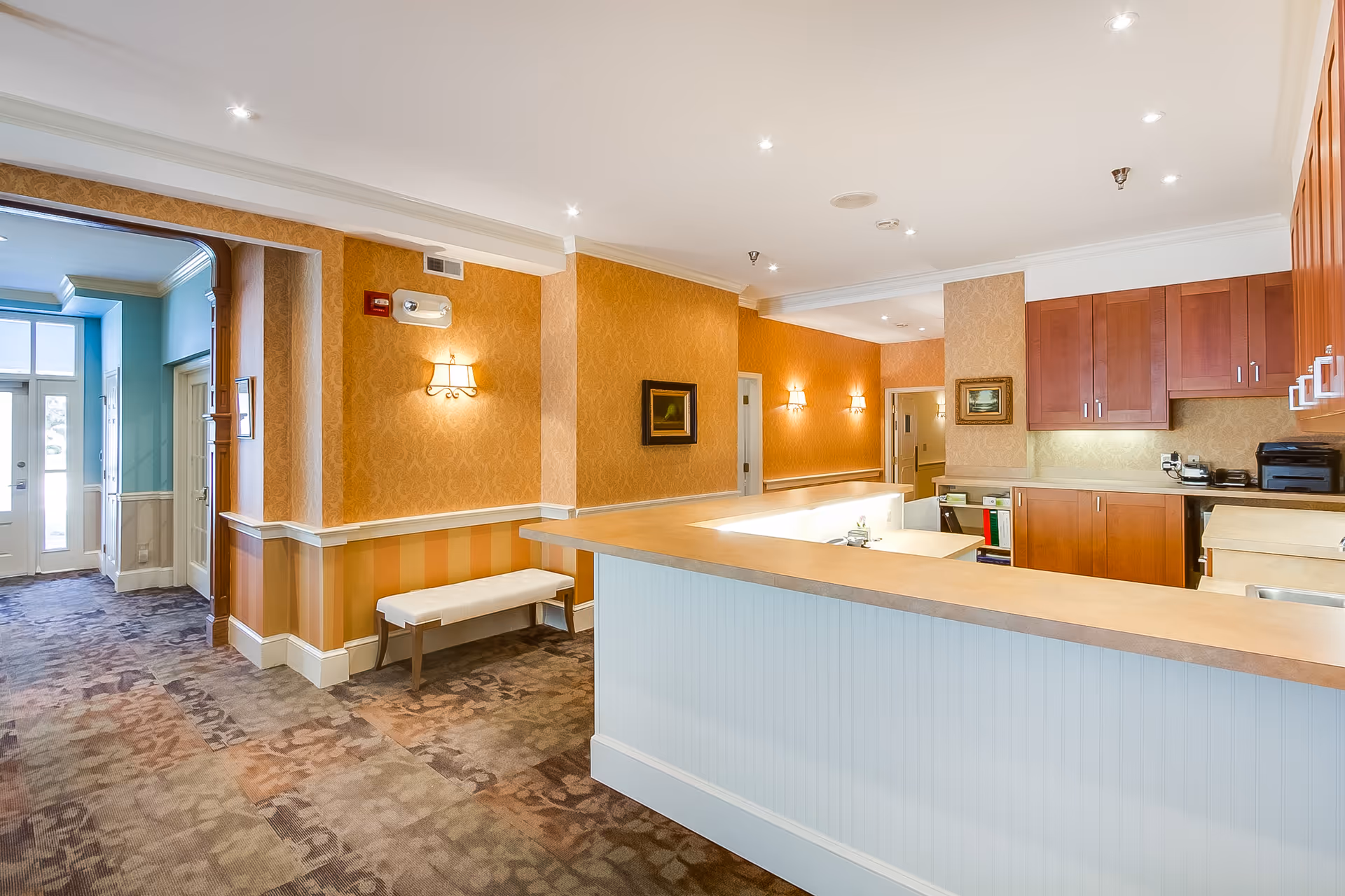 Interior view of a senior living facility reception area with a wooden counter, warm orange patterned wallpaper, wall sconces, framed paintings, and a bench along the wall. The area has carpeted floors and recessed ceiling lights. To the left, there is a hallway with blue walls and a door leading outside.