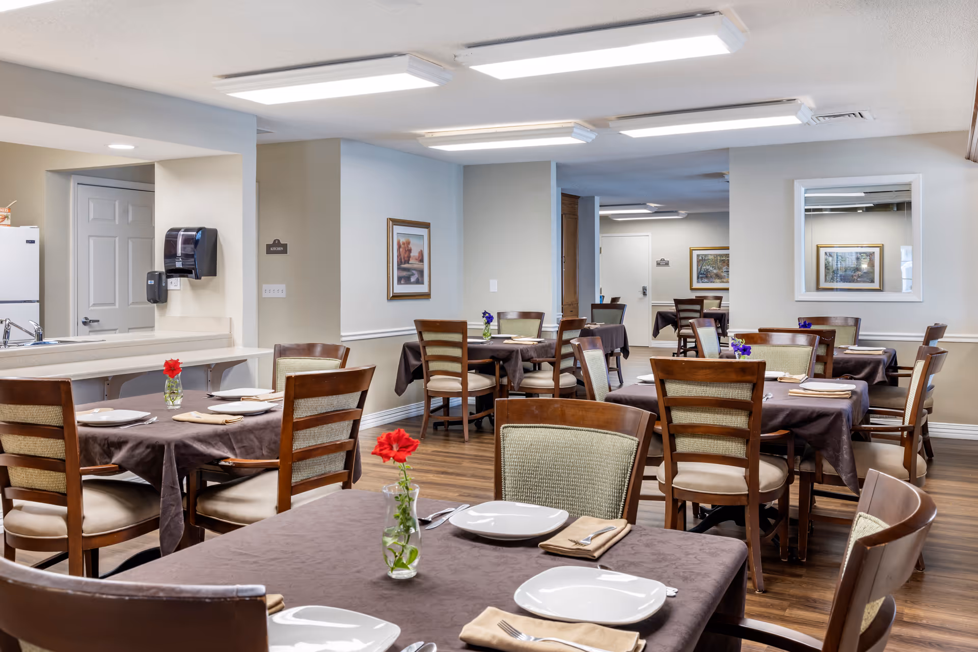 A dining room in a senior living facility with multiple tables covered in brown tablecloths, each set with white plates, beige napkins, and silverware. Small vases with red and purple flowers are placed on the tables. The room has wooden chairs with cushioned seats and backs, wood flooring, and neutral-colored walls decorated with framed artwork and a mirror. Fluorescent ceiling lights illuminate the space.