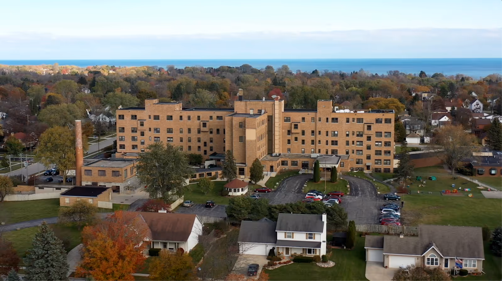 Aerial view of a large multi-story brick building surrounded by trees and residential houses, with a parking lot in front and a body of water visible in the background under a clear sky.