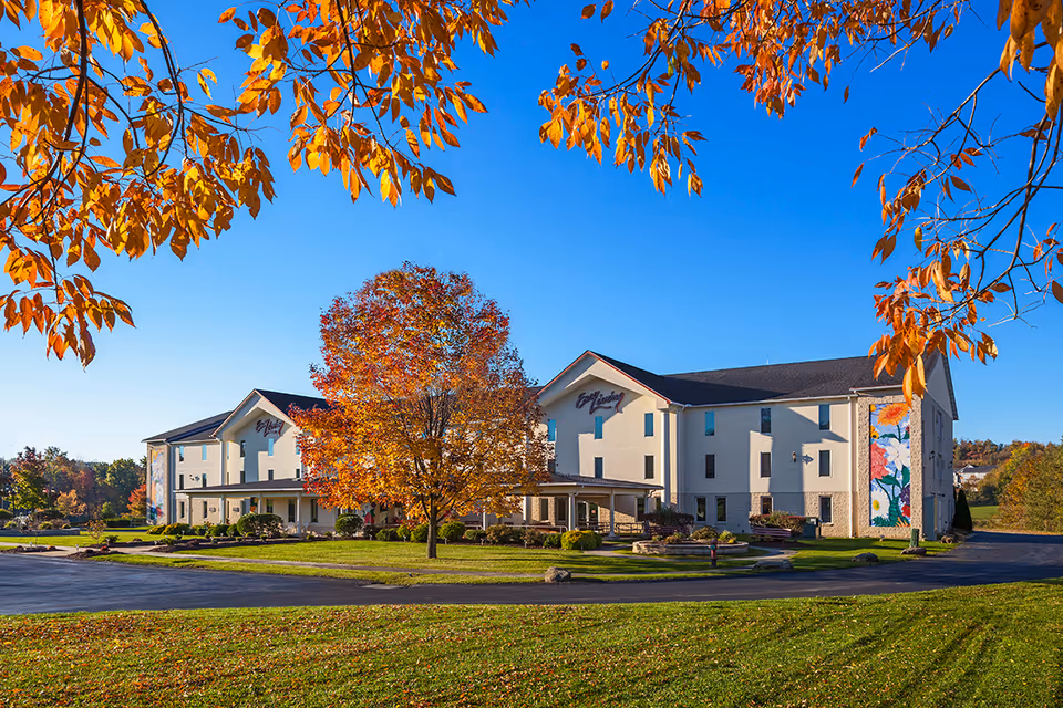 Exterior view of Easy Living Country Estates facility on a clear day with autumn foliage. The building is a two-story structure with light-colored walls and a dark roof, surrounded by green grass and trees with orange and yellow leaves.