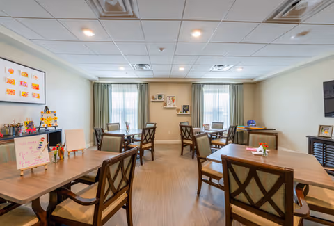 A well-lit room with several wooden tables and chairs arranged neatly, decorated with small flower vases on the tables. The room has two large windows with curtains, framed pictures on the walls, and a whiteboard with colorful writing. The ceiling has recessed lighting and air vents.