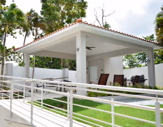 Outdoor covered patio area with a tiled roof, ceiling fan, and seating including chairs and a table, surrounded by green grass and trees in the background.