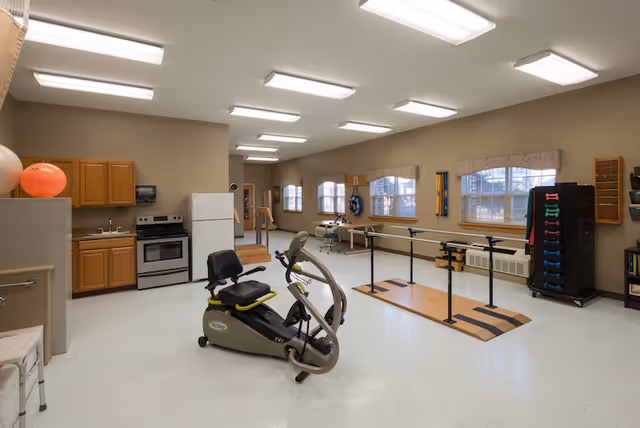 A spacious rehabilitation or physical therapy room with exercise equipment including a recumbent exercise bike, parallel bars for walking practice, and a rack of colorful hand weights. The room has beige walls, white ceiling lights, and several windows with valances allowing natural light. There is a small kitchenette area with wooden cabinets, a refrigerator, and a stove on one side.