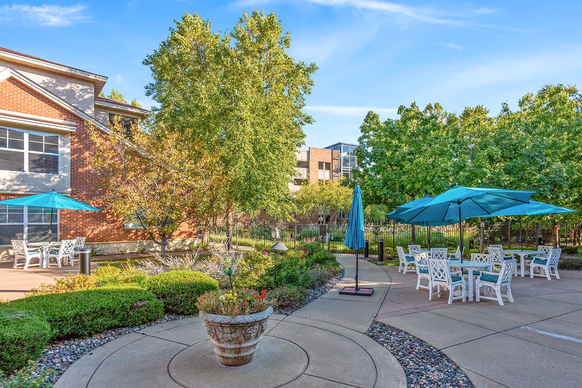 Outdoor courtyard patio with white tables and teal umbrellas surrounded by landscaping and a brick senior living building.